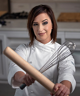 A photo of Anne Lanute holding a wooden rolling pin and metal whisk.